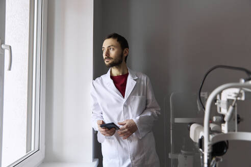 Nachdenklicher Arzt mit Smartphone, der an der Wand in der Nähe des Fensters einer Klinik lehnt - SANF00076