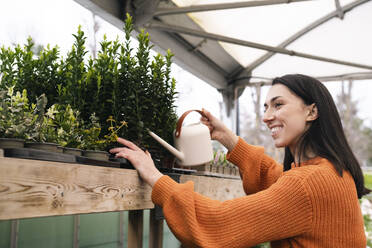 Happy young woman with can watering plants in garden center - AMWF01220