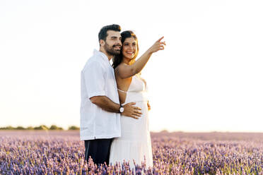 Glückliche Frau, die in die Ferne zeigt, steht mit einem Mann in einem Lavendelfeld bei Sonnenuntergang - JJF00383