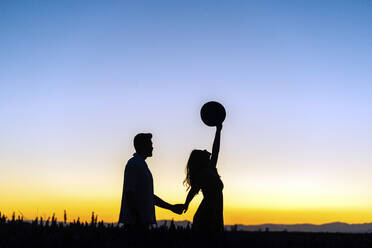 Silhouette man holding hand of girlfriend lifting hat in field at sunset - JJF00420