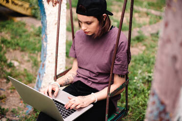 Young female student using laptop sitting on a swing - CAVF96663