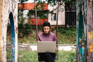 Serious young female student using laptop sitting on a swing in summer - CAVF96665
