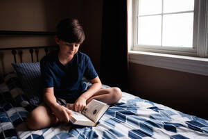 Kleiner Junge liest ruhig ein Buch auf seinem Bett in seinem Schlafzimmer. - CAVF96669