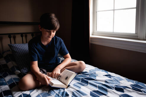 Young boy quietly reading a book on his bed in his bedroom. - CAVF96669