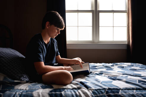 Young boy quietly reading a book on his bed in his bedroom. - CAVF96670