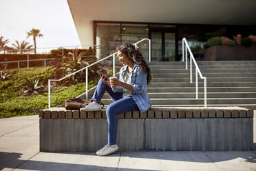 Woman with coffee cup using mobile phone sitting on bench - CAVF96693