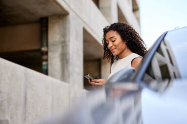 Junge Frau, die in einem Elektroauto eine SMS auf ihrem Smartphone schreibt - CAVF96712