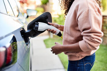 Mittelteil einer jungen Frau, die einen elektrischen Stecker an einem Auto im Hinterhof hält - CAVF96720