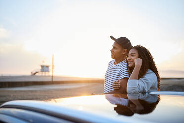 Smiling heterosexual couple by electric car enjoying sunset together - CAVF96724