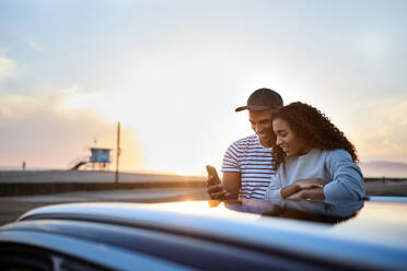 Smiling heterosexual couple using smart phone by car during sunset - CAVF96728