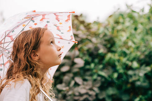 Kind hält einen Regenschirm und schaut bei Regen in den Himmel - CAVF96750