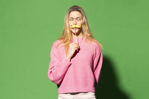 Blonde Frau, die einen Schnurrbart imitiert, vor grünem Hintergrund - MIKF00156