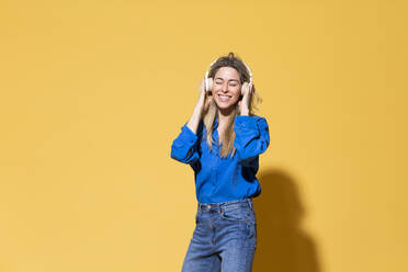 Happy woman enjoying listening music through wireless headphones against yellow background - MIKF00177