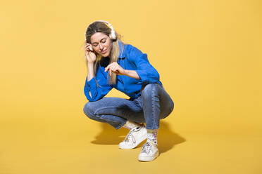 Woman listening to music and crouching against yellow background - MIKF00181