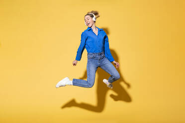 Woman jumping and listening to music against yellow background - MIKF00183