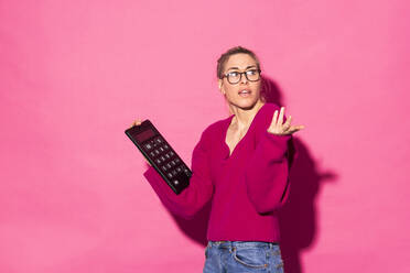 Frau gestikuliert und hält Taschenrechner vor rosa Hintergrund - MIKF00204