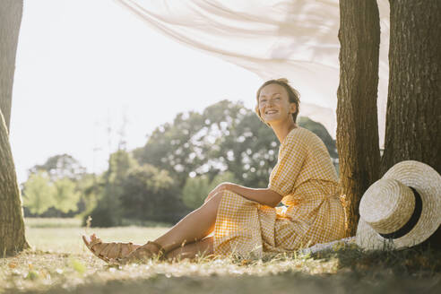 Lächelnde Frau sitzt an einem Baumstamm im Park - NDEF00407