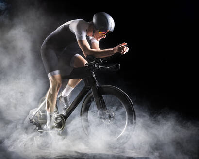 Radfahrer in Sportkleidung auf Turbotrainer sitzend vor schwarzem Hintergrund - STSF03703