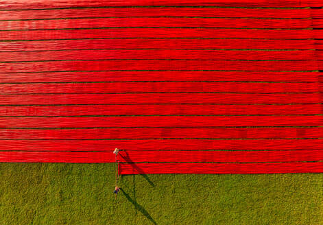 Aerial view of people working in a field stretching red cotton fabric rolls in Narsingdi, Dhaka, Bangladesh. - AAEF17356