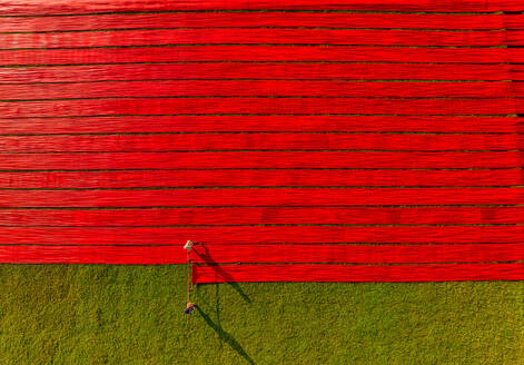 Luftaufnahme von Menschen, die auf einem Feld arbeiten und rote Baumwollstoffrollen in Narsingdi, Dhaka, Bangladesch, spannen. - AAEF17356