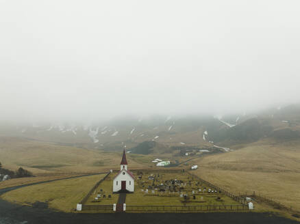 Luftaufnahme der Reyniskirkja-Kirche und des nahe gelegenen Friedhofs, umgeben von Nebel und verschneiten Bergen im Hintergrund in Island. - AAEF17377