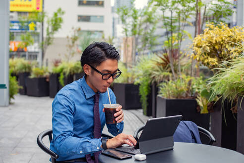 Ein junger Geschäftsmann in der Stadt, der unterwegs ist, ein Mann, der an einem Cafétisch im Freien sitzt, einen Laptop benutzt und mit einem Strohhalm an einem Erfrischungsgetränk nippt. - MINF16597