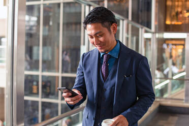 Ein junger Geschäftsmann in einem blauen Anzug in einer Stadt, der auf sein Handy-Display schaut, eine SMS schreibt oder eine Nachricht liest. - MINF16608