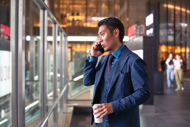 Ein junger Geschäftsmann im blauen Anzug ist in der Innenstadt unterwegs und telefoniert mit seinem Mobiltelefon. - MINF16609