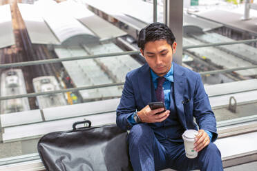 Ein junger Geschäftsmann in einem blauen Anzug in einer Stadt, der auf sein Handy-Display schaut, eine SMS schreibt oder eine Nachricht liest. - MINF16611