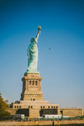 Malerische Seitenansicht der Freiheitsstatue am Ufer in der Nähe von plätscherndem Wasser gegen wolkenlosen blauen Himmel in New York City - ADSF43257