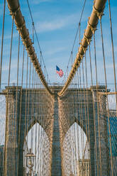 From below of Brooklyn Bridge with USA flag on top against cloudy blue sky in New York City - ADSF43267