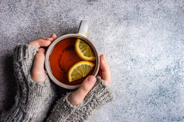 Kid hands in mittens hold cup of tea with lemon on grey concrete background - ADSF43289