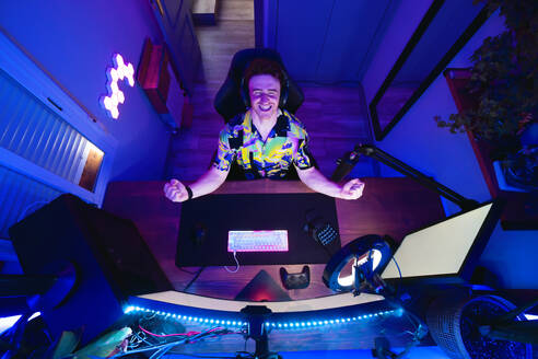 Draufsicht auf einen begeisterten jungen Mann mit Kopfhörern, der mit geballten Fäusten am Tisch sitzt und seinen Erfolg in einem Online-Spiel in einem dunklen Raum mit Neonbeleuchtung feiert - ADSF43305
