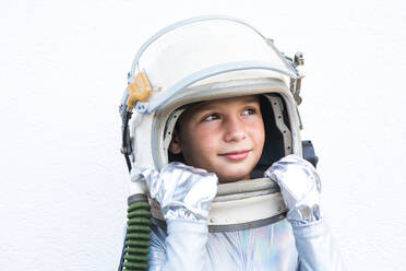 Isolated preteen child in silver space suit touching helmet and looking up while standing against white background - ADSF43312