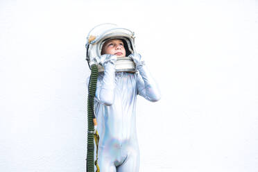 Isolated preteen child in silver space suit touching helmet and looking up while standing against white background - ADSF43313