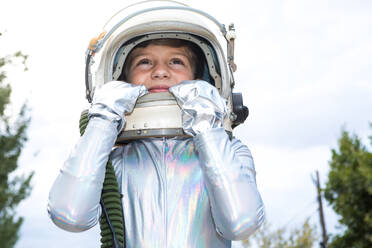 Preteen kid in silver space suit touching helmet and looking up while standing against green trees - ADSF43314