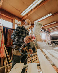 Side view of focused mature male carpenter in protective mask and goggles using electric paint sprayer while varnishing wooden plank - ADSF43316