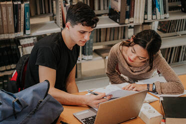 Konzentrierte Teenager-Studenten in legerer Kleidung, die an einem Holztisch mit Laptop sitzen und gemeinsam an einem Universitätsprojekt arbeiten, während sie in der Bibliothek lernen - ADSF43321