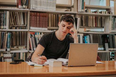 Konzentrierter männlicher Teenager-Student in Freizeitkleidung, der an einem Holztisch mit Laptop sitzt und Notizen schreibt, während er sich in der Bibliothek auf eine Prüfung vorbereitet - ADSF43323