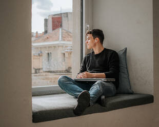 Full body of pensive young male student in casual clothes sitting on windowsill with laptop and looking at window while listening to music in earphones - ADSF43326