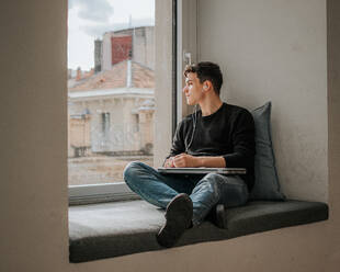 Ganzkörperansicht eines nachdenklichen jungen männlichen Studenten in Freizeitkleidung, der mit seinem Laptop auf der Fensterbank sitzt und zum Fenster schaut, während er mit Kopfhörern Musik hört - ADSF43326