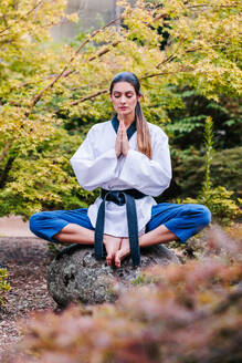 Ganzkörperdarstellung einer ruhigen jungen barfüßigen Taekwondo-Meisterin im Kimono mit schwarzem Gürtel, die während der Meditation auf einem Stein in Lotosstellung sitzt - ADSF43350