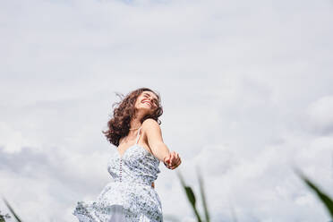 Low angle of happy young female in white summer dress smiling and enjoying sunny day while dancing on grassy lawn - ADSF43352