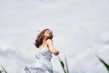 Niedriger Winkel von glücklichen jungen Frau in weißem Sommerkleid lächelnd und genießen sonnigen Tag beim Tanzen auf grasbewachsenen Rasen - ADSF43352