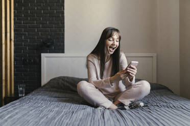 Überrascht junge Frau mit Handy im Schneidersitz auf dem Bett im Schlafzimmer - ANNF00037