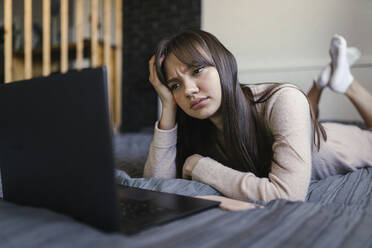 Junge Frau mit Kopf in der Hand, die einen Laptop auf dem Bett zu Hause benutzt - ANNF00042