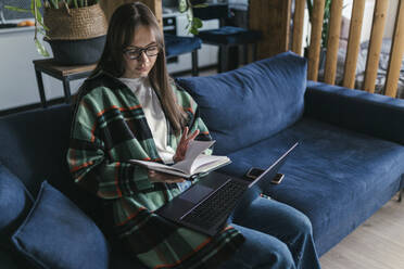 Junge Frau liest ein Buch mit Laptop auf dem Sofa zu Hause - ANNF00068