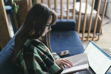 Junge Frau mit Laptop, die zu Hause ein Buch liest - ANNF00069