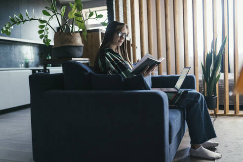 Junge Frau sitzt mit Laptop und liest ein Buch auf dem Sofa zu Hause - ANNF00071