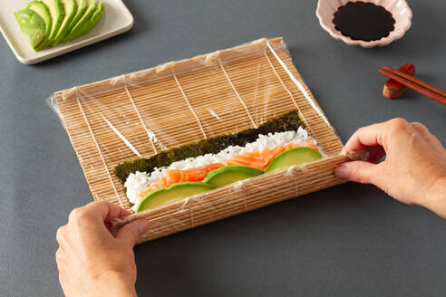 From above of crop anonymous person making sushi consisting of seaweed rice salmon and avocado with bamboo rolling mat on table with chopsticks and soy sauce - ADSF43353